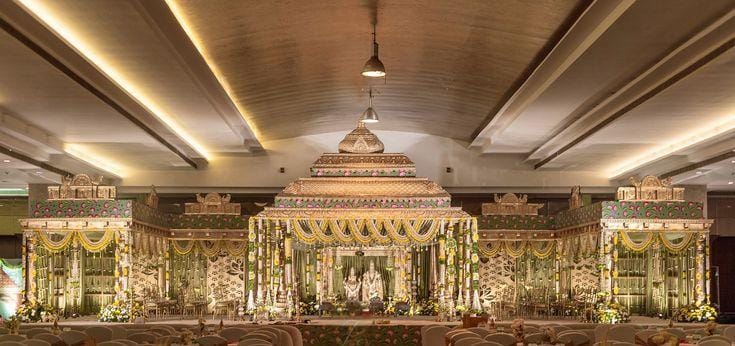 Indoor wedding mandap with carved gold gopuram arches and hanging floral strings by RAW Group